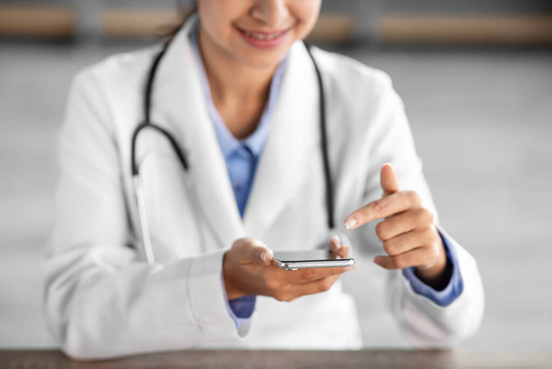 Cropped smiling young hindu lady doctor in white coat with stethoscope surfing in internet on phone - Photo, Image