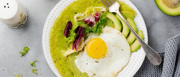Spinach green crepes pancakes with fried egg, avocado and leaves of mix of salad on ceramic plate on gray concrete background. Concept of healthy breakfast. Selective focus. Top view. Copy space. - Foto, Bild