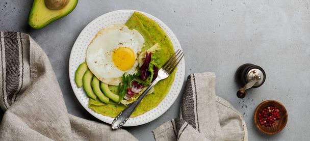 Spinach green crepes pancakes with fried egg, avocado and leaves of mix of salad on ceramic plate on gray concrete background. Concept of healthy breakfast. Selective focus. Top view. Copy space. - Фото, изображение