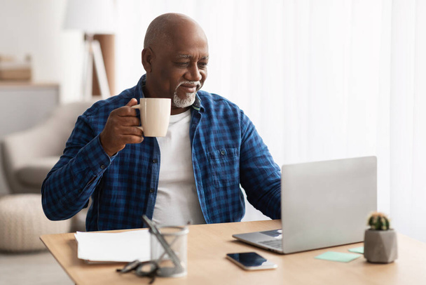 Афроамериканский старший бизнесмен пьет кофе с помощью ноутбука на рабочем месте - Фото, изображение