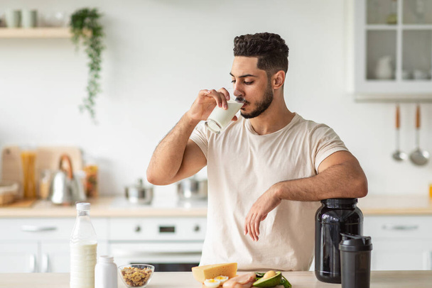 Athletic young Arab man drinking protein shake or milk, standing near table with healthy products at kitchen - Foto, Imagem