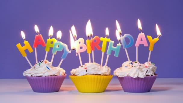 Happy Birthday Inscriptie Gemaakt van Brandende Kleurrijke Kaarsen op Lekkere Cupcakes geïsoleerd op paarse achtergrond. Gelukkige verjaardag video banner, slow motion - Video