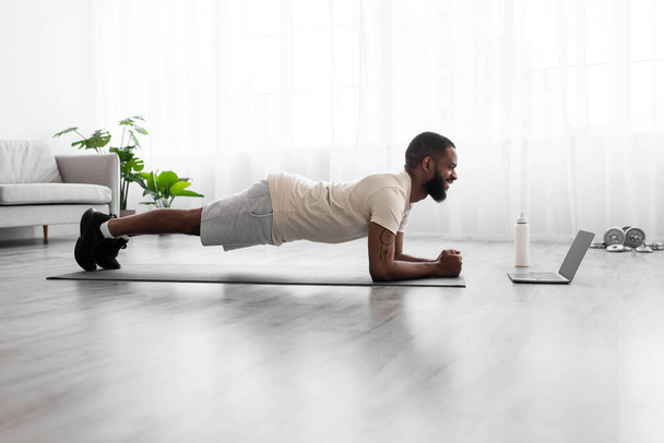 Happy young black man with beard in white sportswear do plank, watch video on laptop in room - Photo, Image