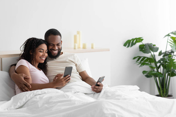 Smiling young african american husband and wife hugging, looking at smartphones on white bed - Foto, imagen