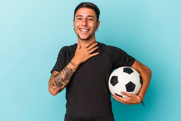 Young man playing soccer holding a ball isolated on blue background  laughs out loudly keeping hand on chest. - 写真・画像