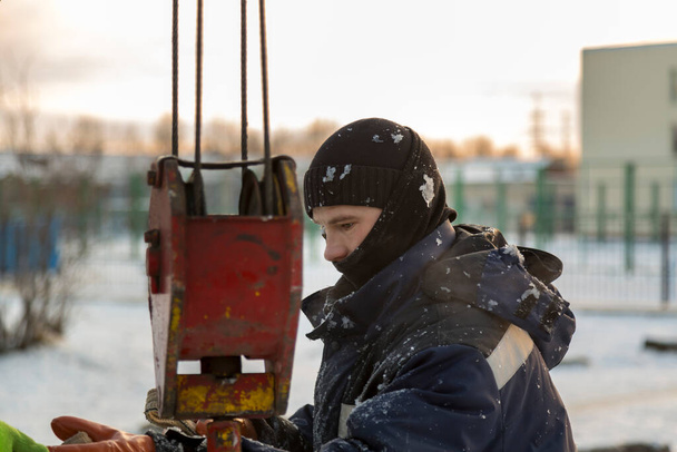 Portret van een arbeider in een blauw jasje met een kap aan de haak van een vrachtwagenkraan bij de bouw van een ijsstad - Foto, afbeelding