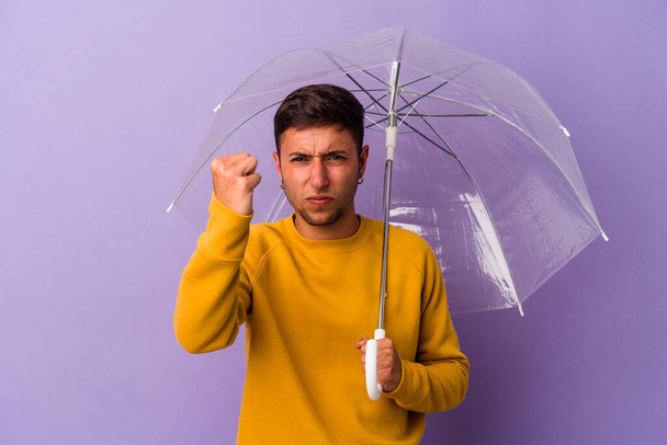 młody biały mężczyzna trzymając parasol odizolowany na fioletowym tle pokazując pięść do kamery, agresywny wyraz twarzy. - Zdjęcie, obraz