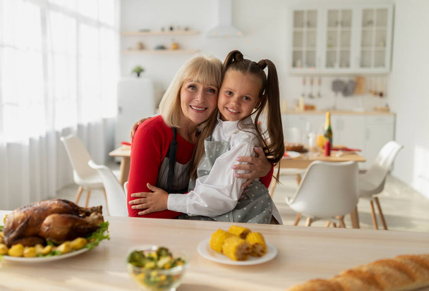Portrét veselé holčičky a její babičky v zástěrách vaření vánoční večeře, pózování a objímání v kuchyni - Fotografie, Obrázek