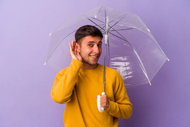 Young caucasian man holding umbrella isolated on purple background  trying to listening a gossip. - Foto, imagen