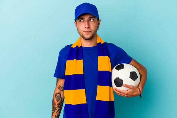 Młody fan piłki nożnej trzyma piłkę odizolowany na niebieskim tle mylić, czuje wątpliwości i niepewność. - Zdjęcie, obraz