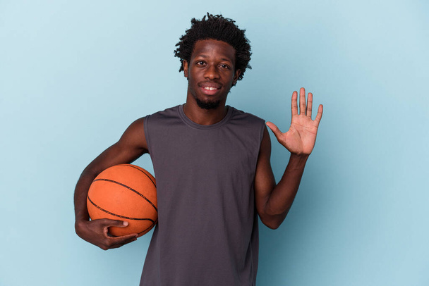 Young african american man playing basketball isolated on blue background smiling cheerful showing number five with fingers. - 写真・画像