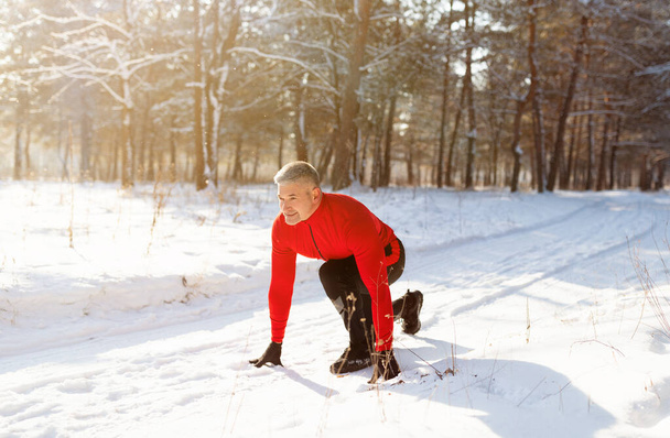 Alegre hombre mayor listo para correr en el camino nevado del campo en el frío día de invierno, longitud completa. Concepto de estilo activo - Foto, imagen