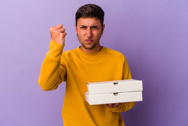 Young caucasian man holding pizzas isolated on purple background  showing fist to camera, aggressive facial expression. - Foto, Imagen