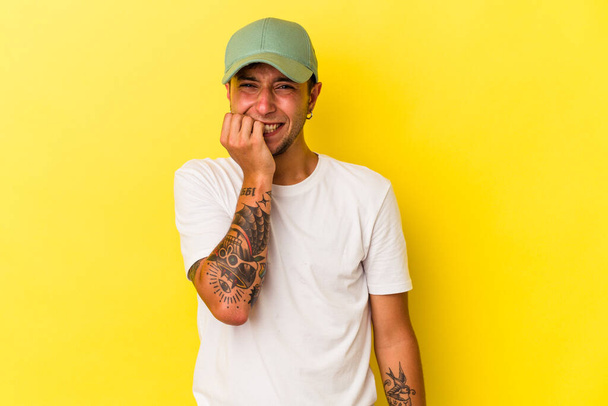 Young caucasian man with tattoos isolated on yellow background  biting fingernails, nervous and very anxious. - Фото, зображення