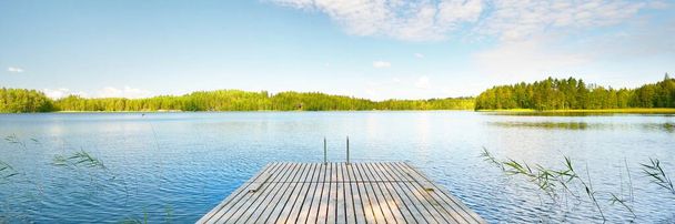 Wooden pier near the river close-up. Evergreen coniferous forest in the background. Clear blue sky, reflections on the water. Idyllic summer landscape. Eco tourism, recreation theme. Finland - Photo, Image