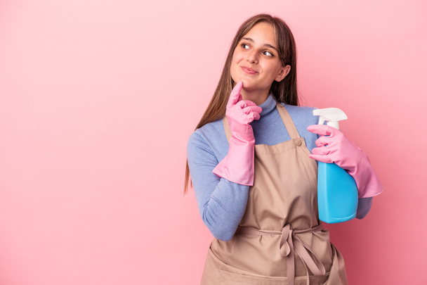Mujer joven limpiadora caucásica sosteniendo aerosol aislado sobre fondo rosa mirando hacia los lados con expresión dudosa y escéptica. - Foto, imagen