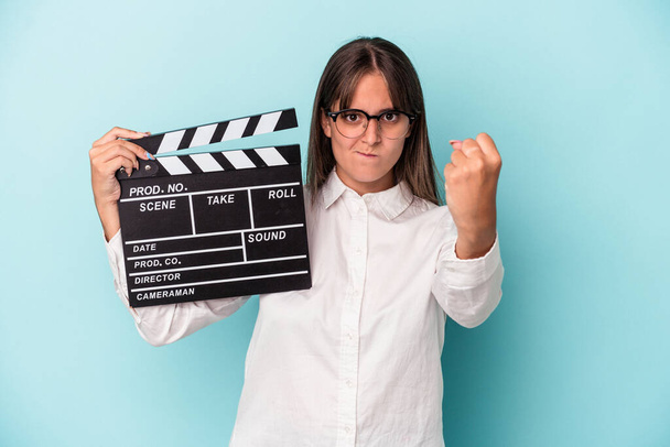 Young caucasian woman holding clapperboard isolated on blue background showing fist to camera, aggressive facial expression. - Фото, зображення