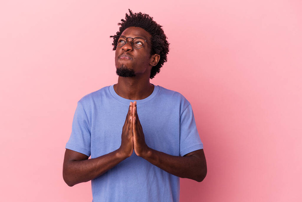 młody Afroamerykanin człowiek odizolowany na różowym tle trzymając się za ręce w modlitwie blisko ust, czuje się pewnie. - Zdjęcie, obraz
