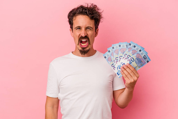 Junger kaukasischer Mann hält Geldscheine isoliert auf rosa Hintergrund und schreit sehr wütend und aggressiv. - Foto, Bild