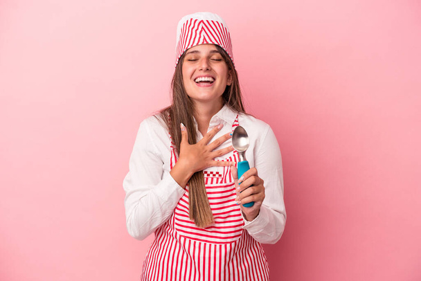 Молода жінка-виробник морозива, що тримає ложку ізольовану на рожевому фоні, голосно сміється, тримаючи руку на грудях
. - Фото, зображення