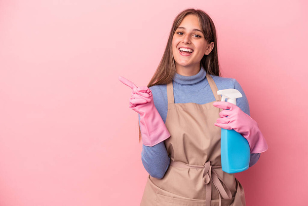 Young caucasian cleaner woman holding spray isolated on pink background smiling and pointing aside, showing something at blank space. - Photo, Image