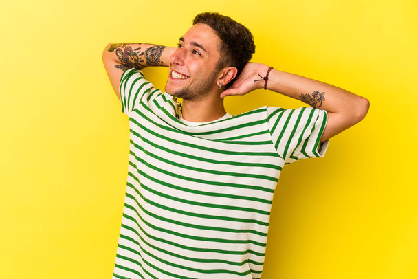 Νεαρός καυκάσιος άνδρας με τατουάζ απομονωμένο σε κίτρινο φόντο αισθάνεται αυτοπεποίθηση, με τα χέρια πίσω από το κεφάλι. - Φωτογραφία, εικόνα