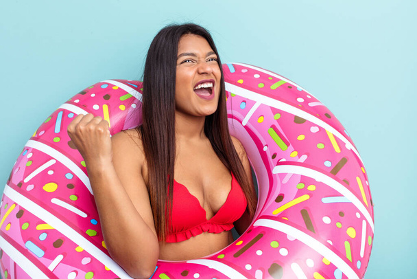 jonge Venezolaanse vrouw houden donut opblaasbaar geïsoleerd op blauwe achtergrond raisen vuist na een overwinning, winnaar concept. - Foto, afbeelding