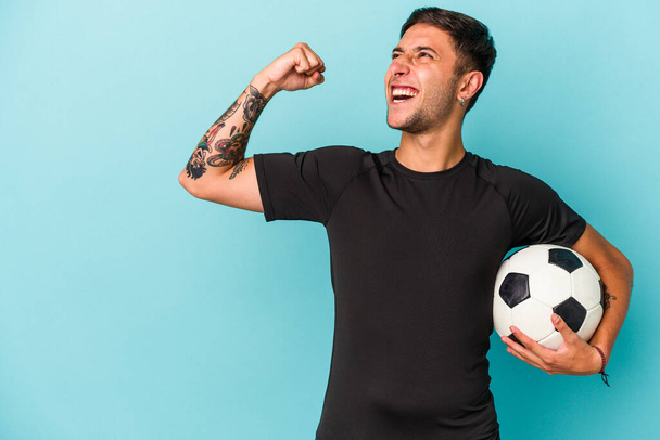 Νεαρός που παίζει ποδόσφαιρο κρατώντας μια μπάλα απομονωμένη σε μπλε φόντο υψώνοντας γροθιά μετά από μια νίκη, νικήτρια ιδέα. - Φωτογραφία, εικόνα