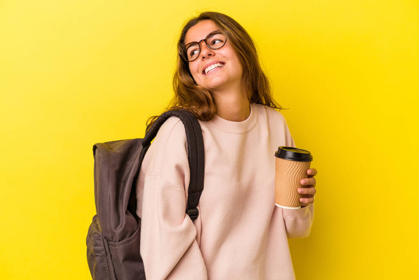 Young caucasian student woman holding coffee isolated on yellow background  looks aside smiling, cheerful and pleasant. - Foto, imagen