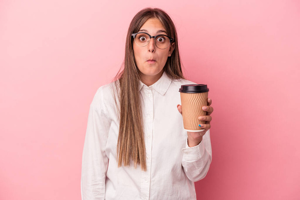 Young business caucasian woman holding a take away isolated on pink background shrugs shoulders and open eyes confused. - Photo, Image