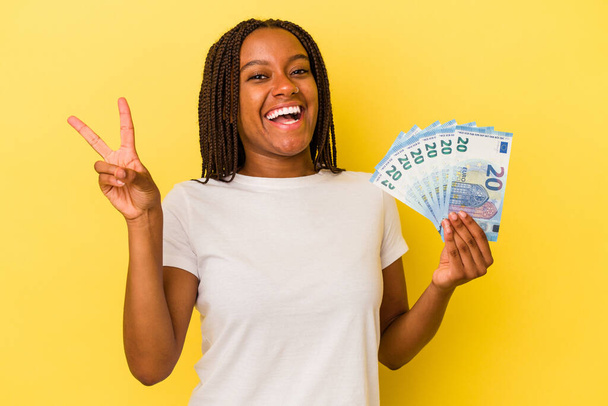 Молодая африканская американка держит банкноты на желтом фоне радостно и беззаботно показывая символ мира с пальцами. - Фото, изображение