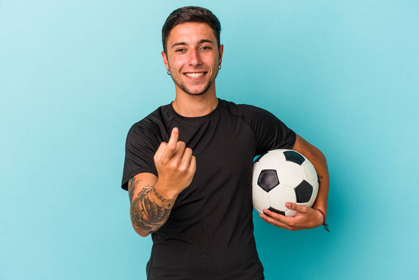 Young man playing soccer holding a ball isolated on blue background  pointing with finger at you as if inviting come closer. - 写真・画像