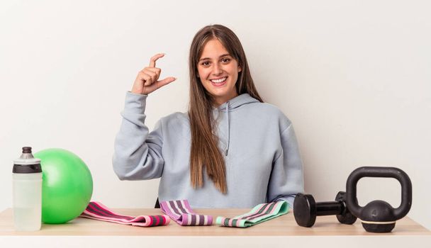 Young caucasian woman sitting at a table with sport equipment isolated on white background holding something little with forefingers, smiling and confident. - Photo, Image