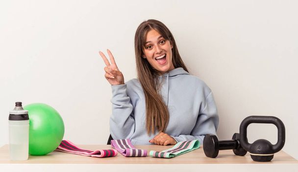 Young caucasian woman sitting at a table with sport equipment isolated on white background joyful and carefree showing a peace symbol with fingers. - Photo, Image