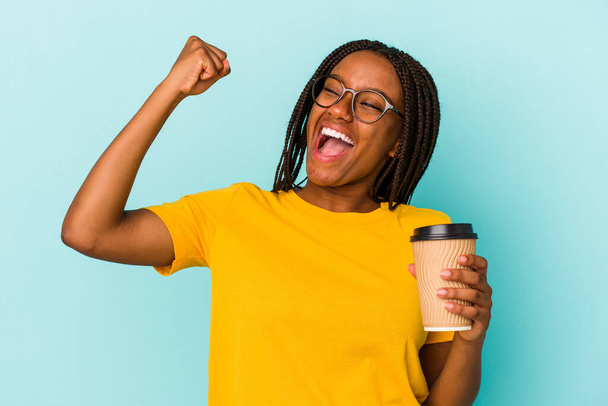 Νεαρή Αφροαμερικανή γυναίκα κρατώντας ένα take away καφέ απομονωμένη σε μπλε φόντο αυξάνοντας γροθιά μετά από μια νίκη, νικητής έννοια. - Φωτογραφία, εικόνα