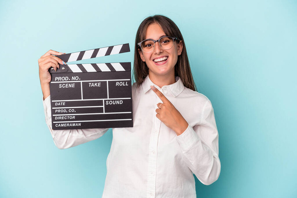 Young caucasian woman holding clapperboard isolated on blue background smiling and pointing aside, showing something at blank space. - Fotografie, Obrázek