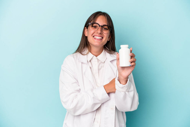 Joven mujer farmacéutica sosteniendo pastillas aisladas sobre fondo azul riendo y divirtiéndose. - Foto, Imagen