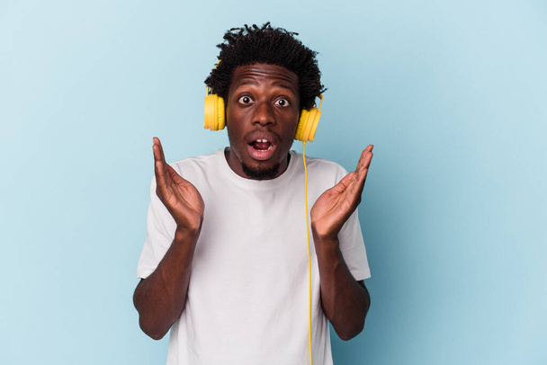 Joven afroamericano escuchando música aislada sobre fondo azul sorprendido y sorprendido. - Foto, imagen