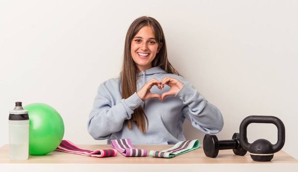 Giovane donna caucasica seduta a un tavolo con attrezzature sportive isolate su sfondo bianco sorridente e mostrando una forma a cuore con le mani. - Foto, immagini