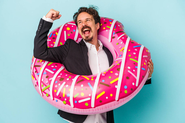 Joven hombre de negocios con donut inflable aislado sobre fondo azul levantando puño después de una victoria, concepto ganador. - Foto, imagen