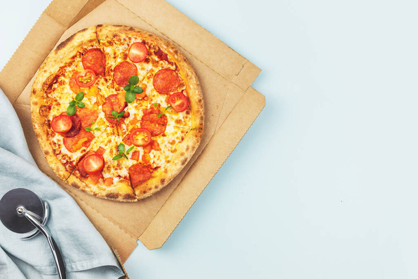 Natural italian big pepperoni pizza with thin dough, salami, mozzarella, cherry tomatoes and basil in a craft box on a blue background with copy space. Mediterranean cuisine. Pizza delivery. Flat lay - Foto, imagen