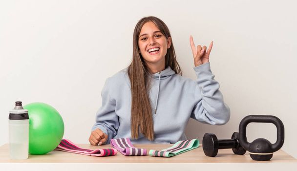 Young caucasian woman sitting at a table with sport equipment isolated on white background showing a horns gesture as a revolution concept. - Photo, Image