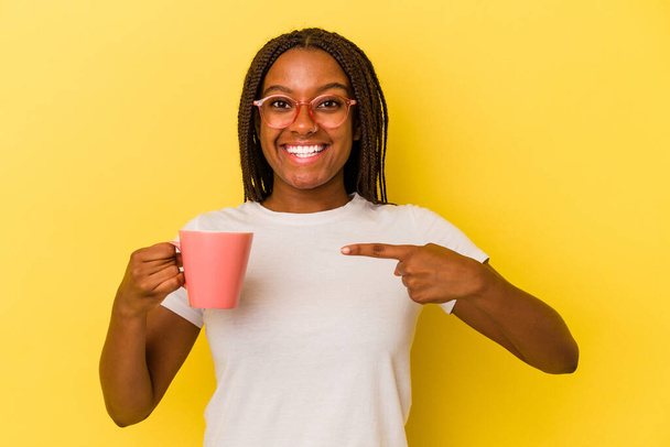 黄色の背景にマグカップを持った若いアフリカ系アメリカ人女性が笑顔で脇を指差して. - 写真・画像