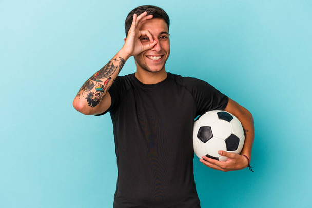 Nuori mies pelaa jalkapalloa pitämällä pallo eristetty sinisellä taustalla innoissaan pitää ok ele silmään. - Valokuva, kuva