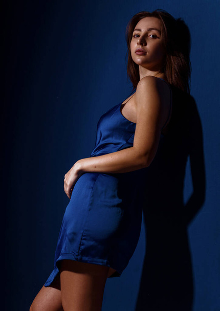 Young and cute brunette woman in blu dress posing on dark blue background free space for text - Foto, imagen