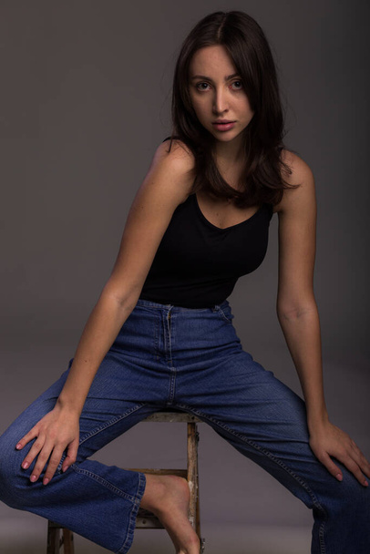 Fashion model sit on chair in a blu jeans and black undershirt barefoot on studio gray background - Foto, Imagem