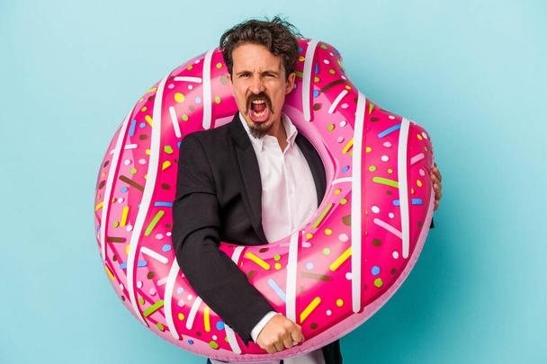 Young business man with inflatable donut isolated on blue background screaming very angry and aggressive. - Foto, imagen