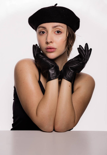 Portrait of gorgeous young fashion model in black beret and leather gloves leaning on the table on white background. Closeup face of young beautiful woman with a healthy clean skin - Φωτογραφία, εικόνα