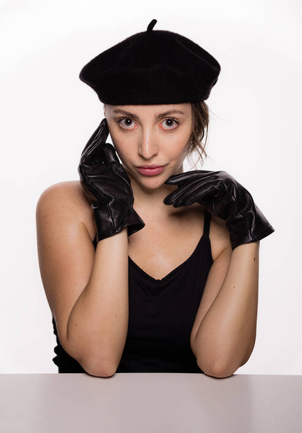 Πορτρέτο του πανέμορφο νεαρό μοντέλο μόδας σε μαύρο μπερέ και δερμάτινα γάντια ακουμπά στο τραπέζι σε λευκό φόντο. Κοντινό πλάνο πρόσωπο της νεαρής όμορφης γυναίκας με υγιές καθαρό δέρμα - Φωτογραφία, εικόνα