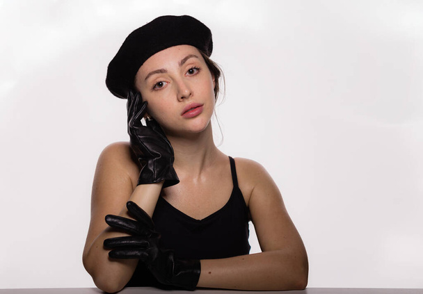 Portrait of gorgeous young fashion model in black beret and leather gloves leaning on the table on white background. Closeup face of young beautiful woman with a healthy clean skin - Fotoğraf, Görsel
